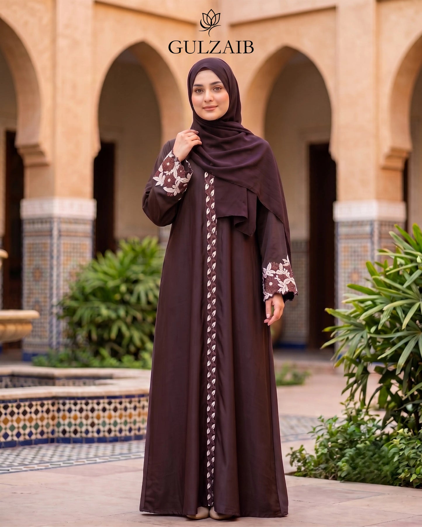A woman wearing a dark brown abaya with floral-patterned sleeves and a matching hijab stands in front of an arched courtyard. The brand name 'GULZAIB' is displayed at the top.