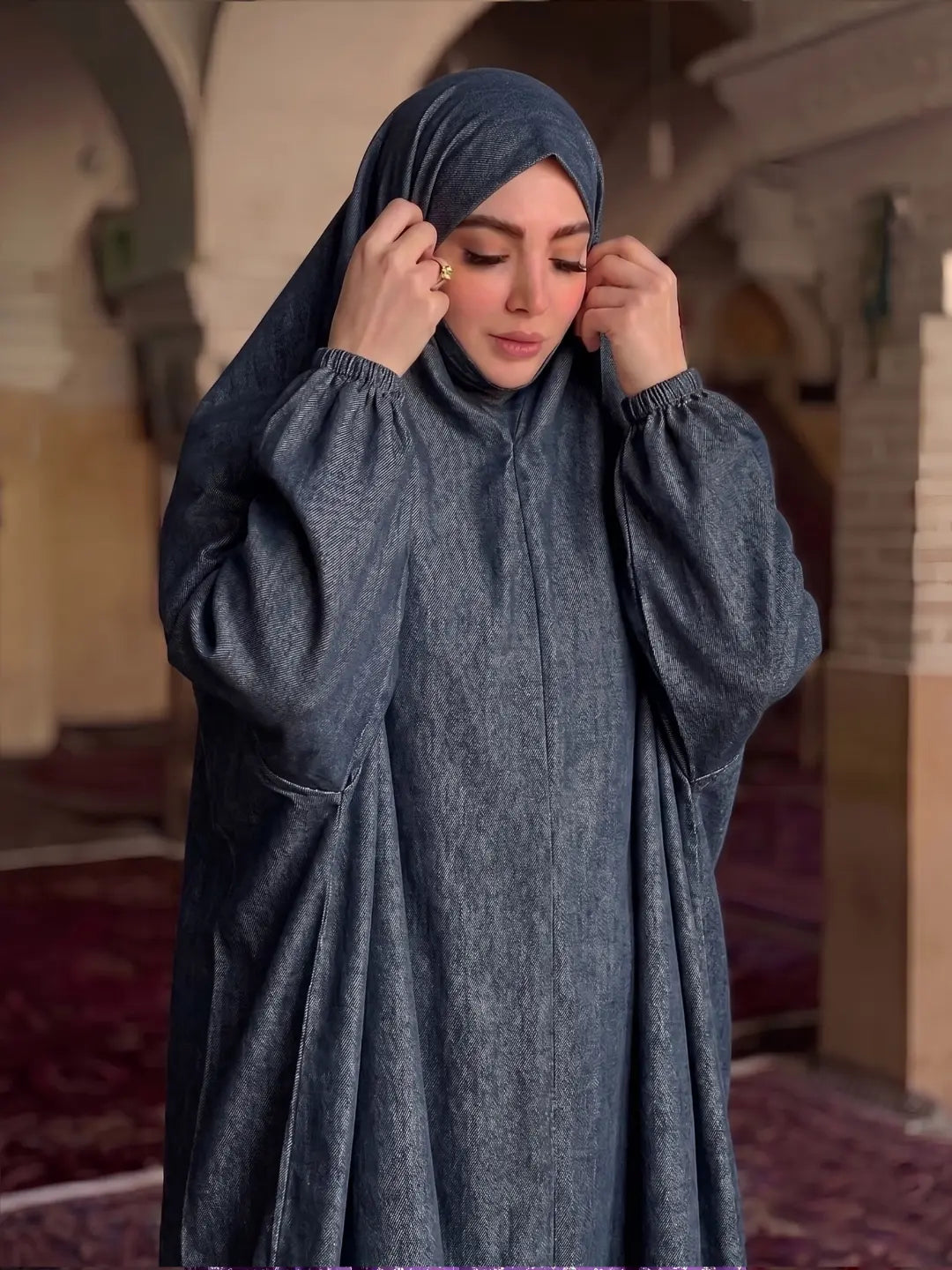 A woman wearing a dark slate blue-gray modest abaya and matching hijab, standing inside a historic building with arches and carpeted floors.