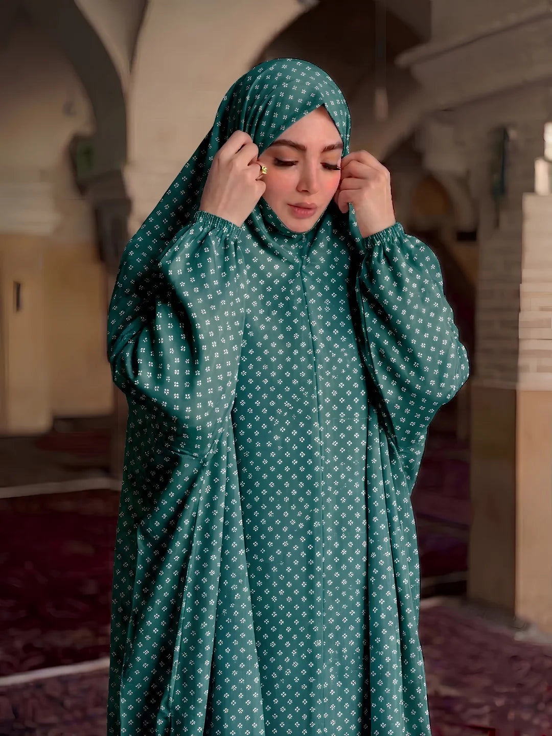 A woman wearing a teal green modest abaya and matching hijab with a small white geometric pattern, standing in a historic corridor with arches.
