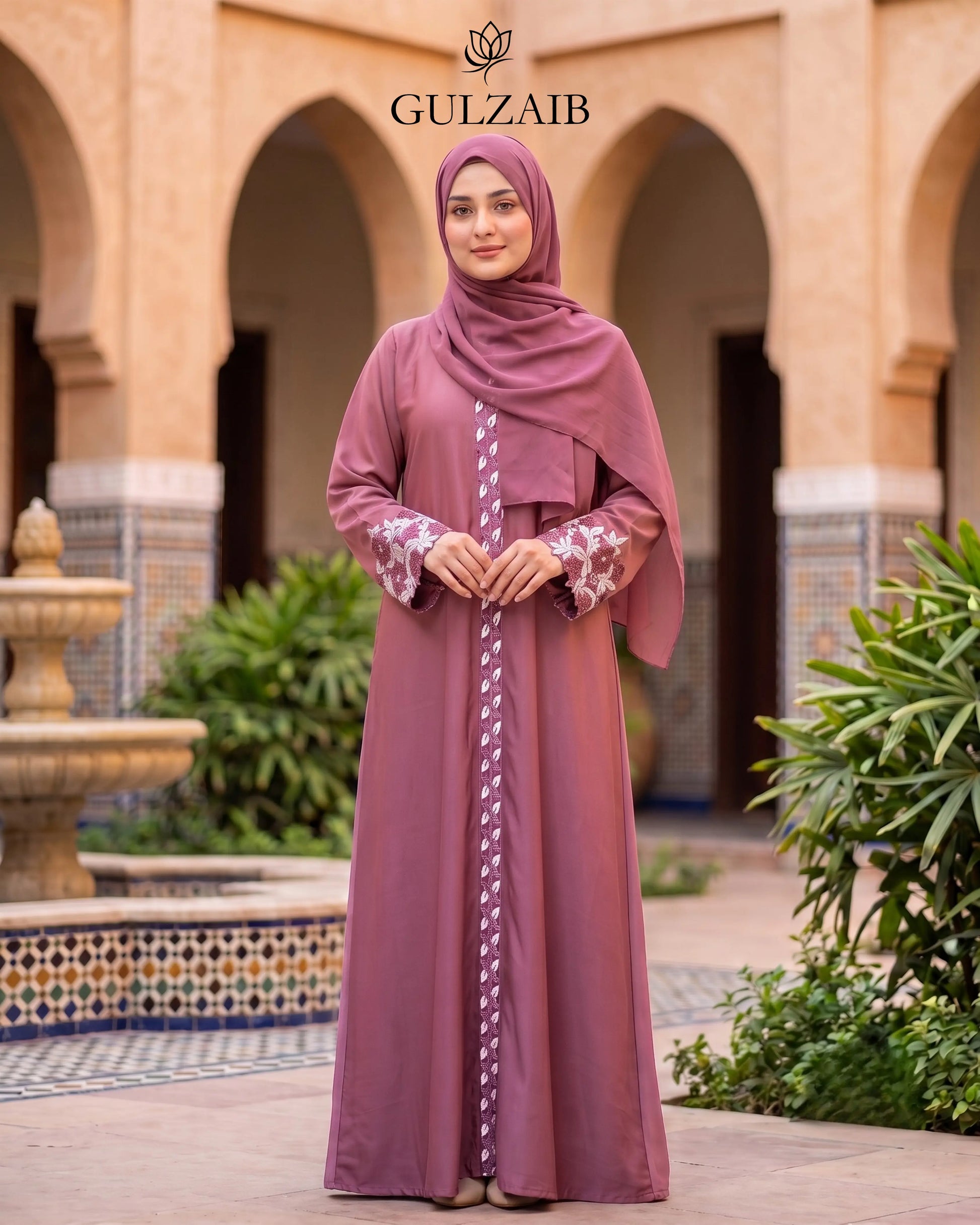 A woman wearing a dusty rose abaya with white floral embroidery and a matching hijab stands in front of a traditional courtyard with arches. The brand name 'GULZAIB' is displayed at the top.