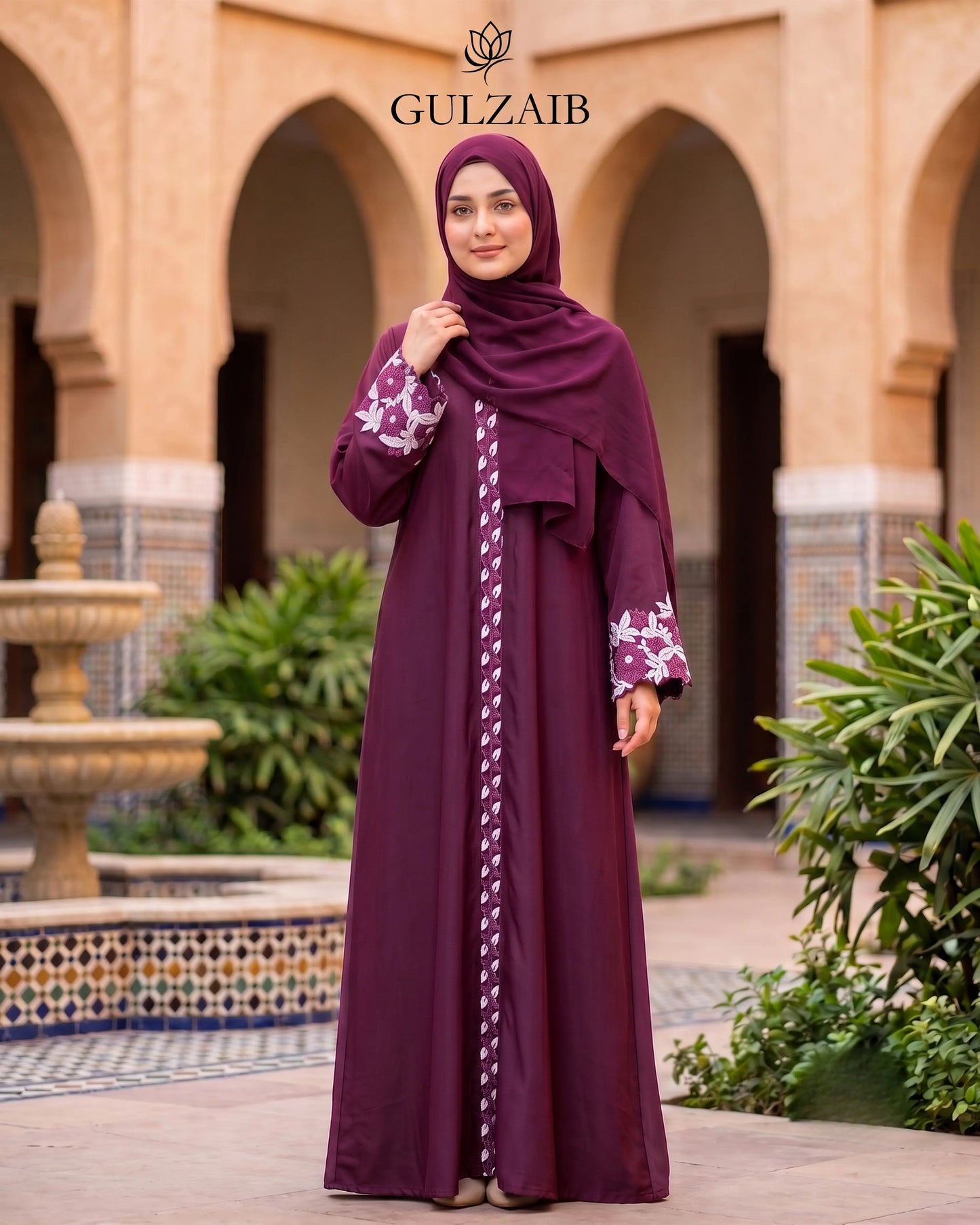 A woman wearing a dark maroon abaya with floral embroidery on the sleeves and a matching hijab, standing in front of a traditional courtyard with arches. The brand name 'GULZAIB' is displayed at the top.
