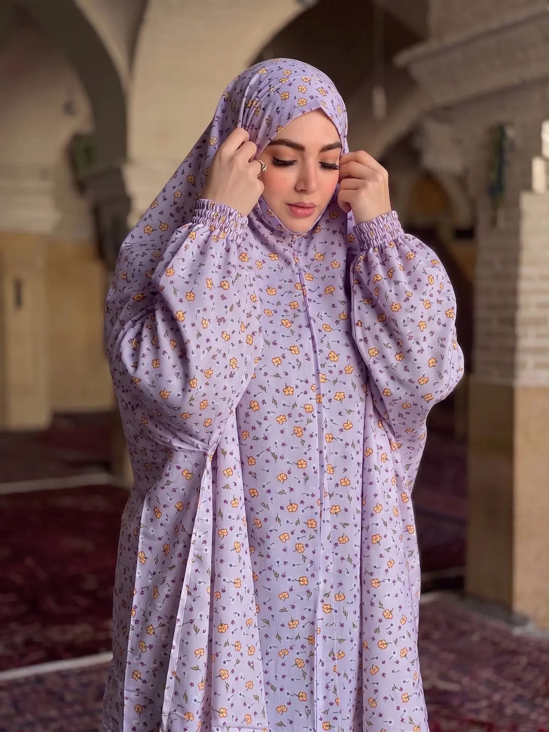A woman wearing a light purple/lavender modest outfit with a matching hijab, featuring a small orange and yellow floral pattern. She is standing inside a mosque or hall with arched ceilings and carpeted floors.