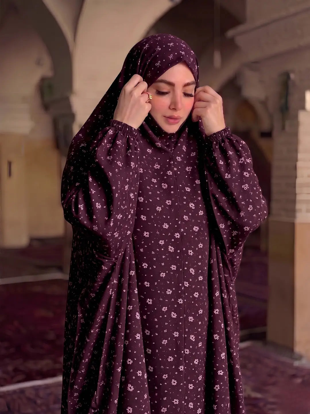 A woman wearing a dark plum maroon modest outfit with a matching hijab and long sleeves, featuring a small white floral pattern. She is standing in a softly lit mosque or hall with arches and carpeted floors.
