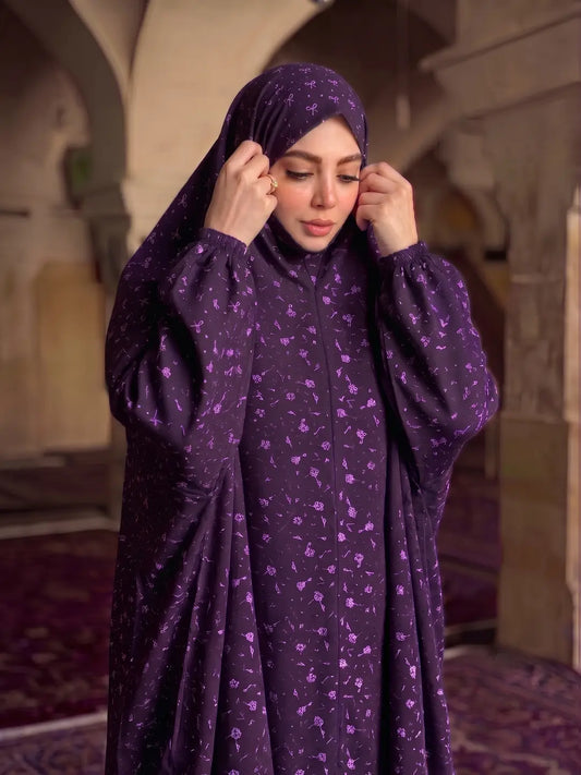 A woman wearing a deep purple modest outfit with a matching patterned hijab, standing in a historic stone corridor with arches.