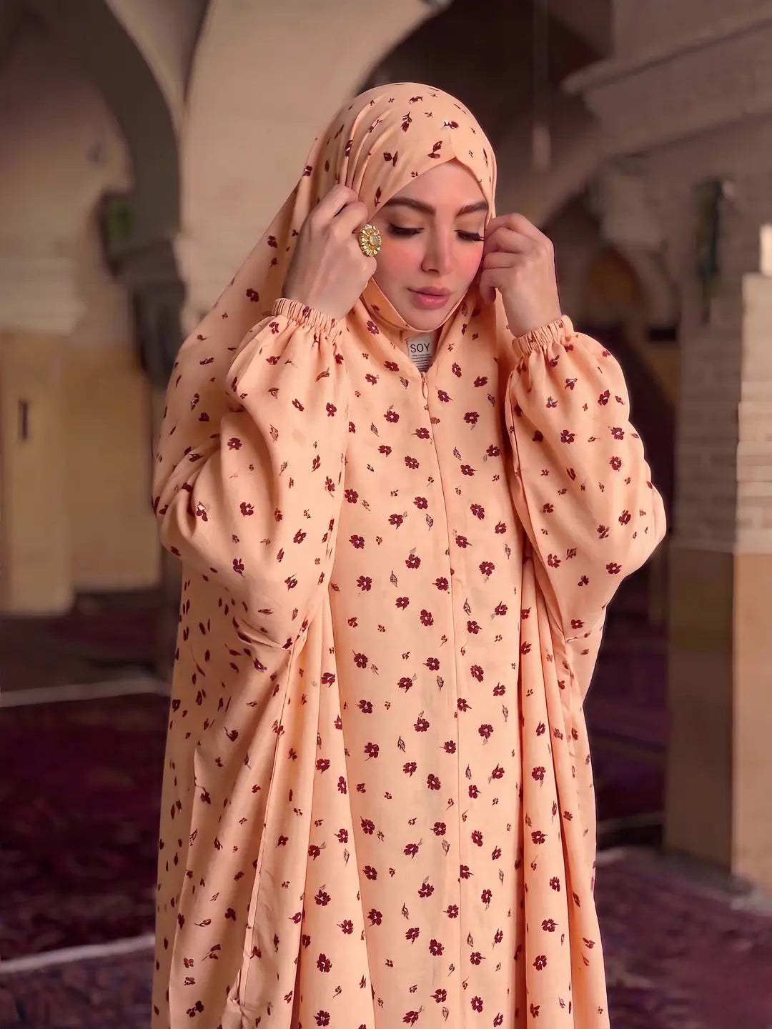 A woman wearing a peach-colored modest outfit with a small red floral pattern, including a long-sleeved abaya and matching hijab, standing in an indoor architectural setting with arches.
