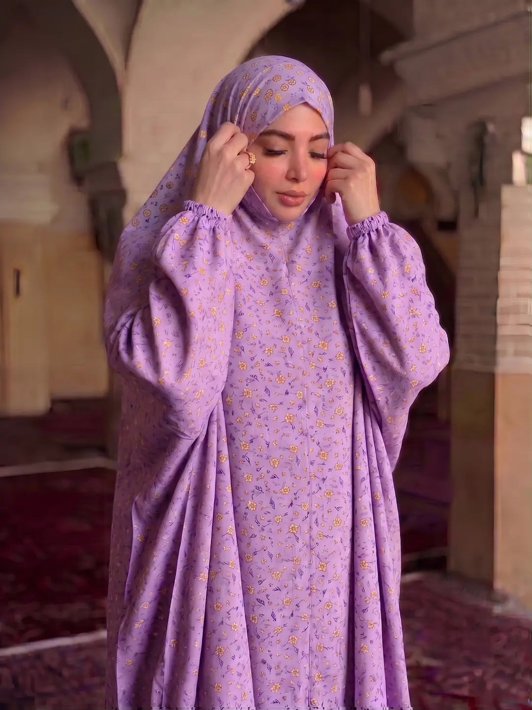 A woman wearing a lavender purple modest outfit with a matching patterned hijab, standing in a historic stone corridor with arches and carpeted floors.