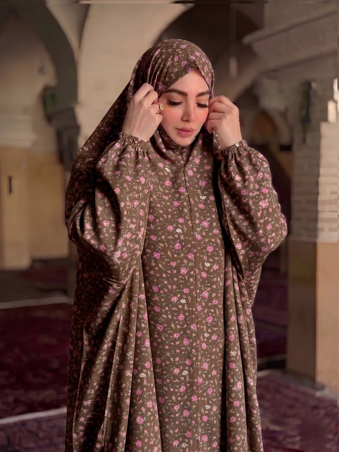 A woman wearing a brown floral hijab and a matching long-sleeved abaya with a small pink and white floral print, standing in a mosque interior with arched corridors and carpeted floors.