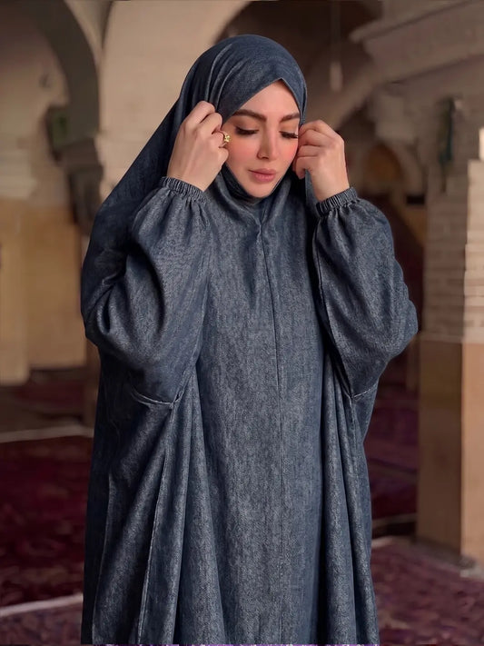 A woman wearing a dark slate blue-gray modest abaya and matching hijab, standing inside a historic building with arches and carpeted floors.