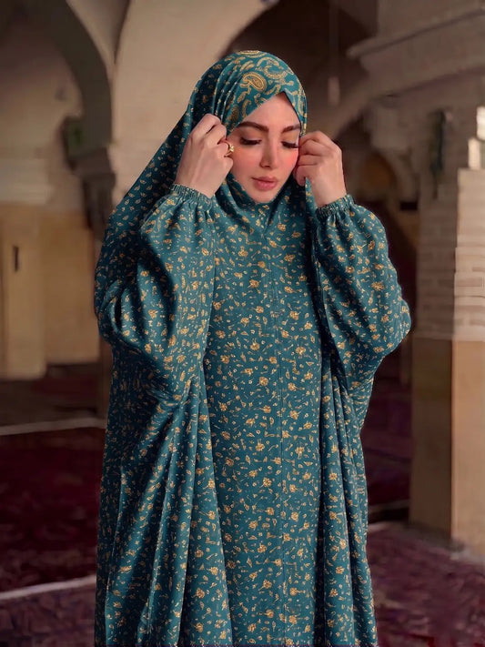 A woman wearing a teal green modest long-sleeve abaya with a small yellow floral pattern and a matching patterned hijab, standing in a historic corridor with arches.
