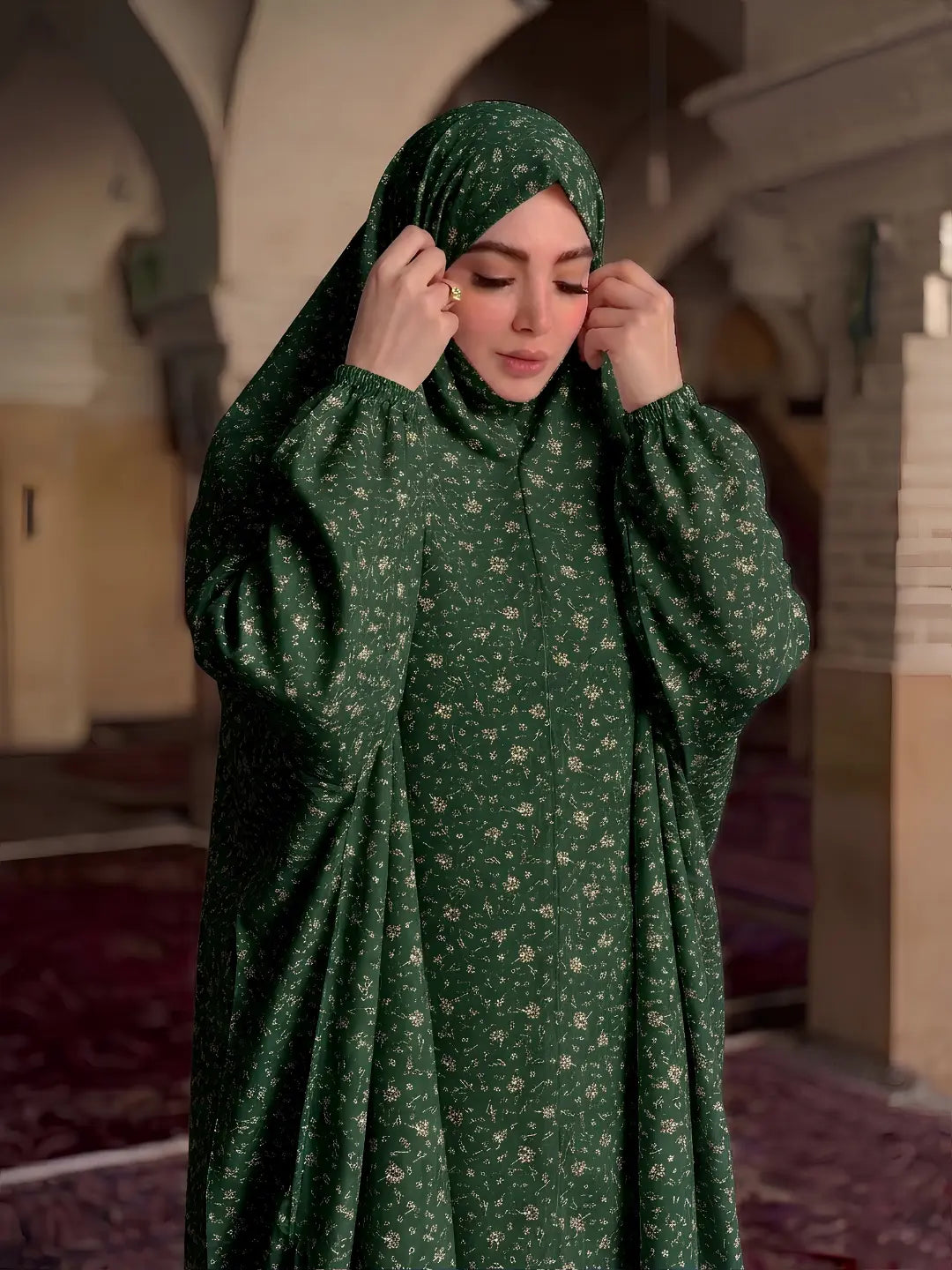A woman wearing a dark green floral abaya and matching hijab, adjusting her headscarf in a softly lit indoor setting with arches and carpeted floors.