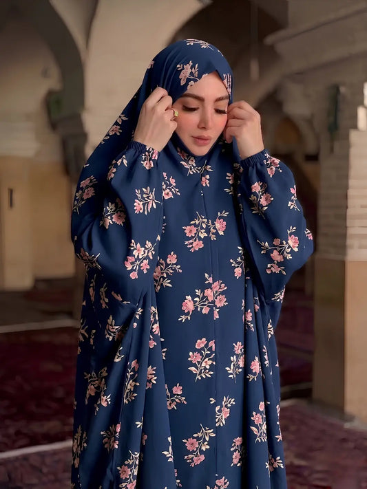 A woman wearing a navy blue modest dress with a pink and beige floral print, paired with a matching navy blue hijab, standing in a historic interior with arches.