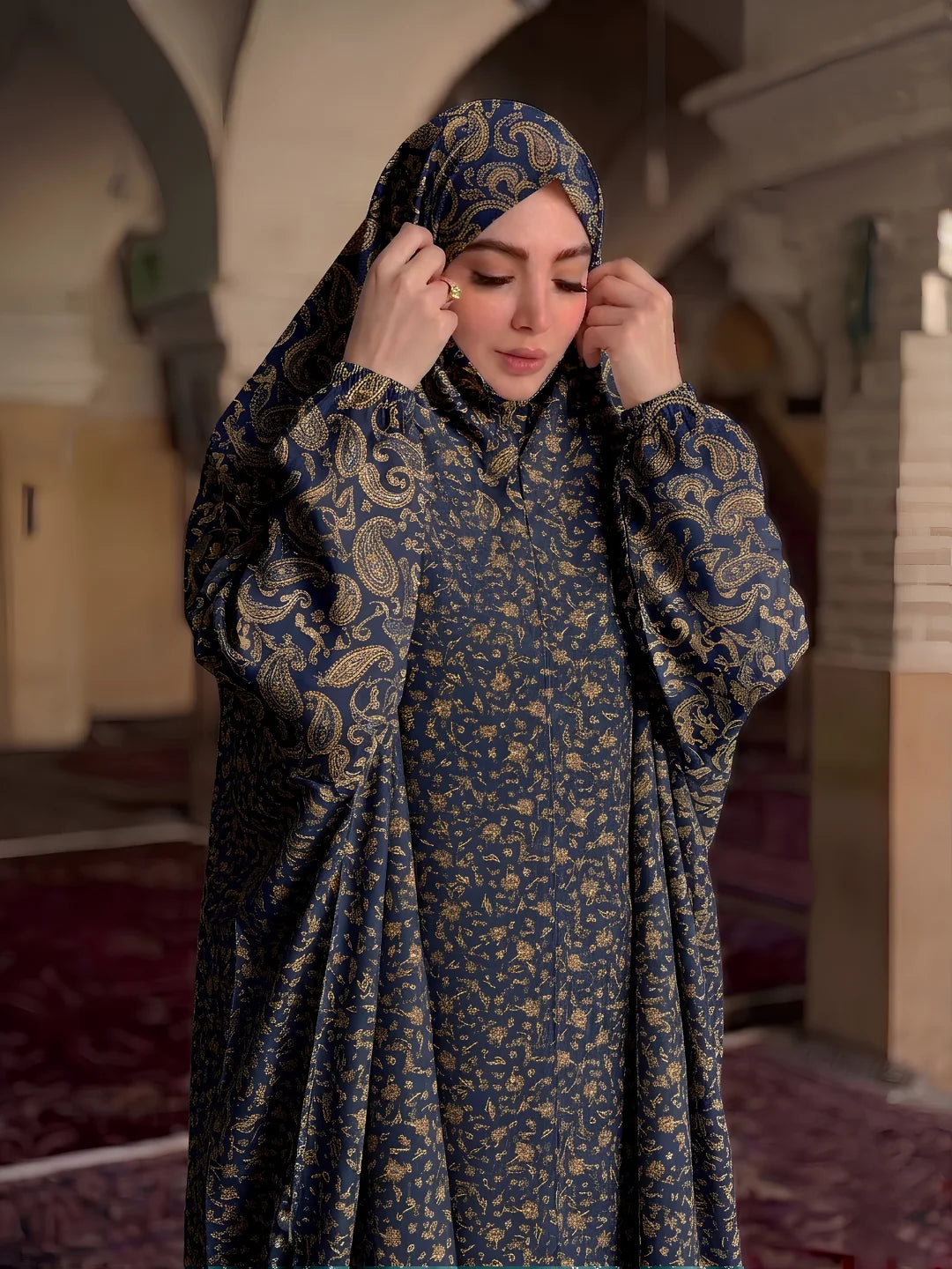 A woman wearing a long-sleeved navy blue and gold patterned abaya with a matching hijab, standing inside a mosque with arched corridors and prayer rugs in the background.