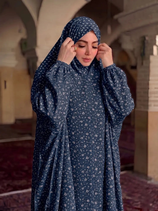 A woman wearing a navy blue modest outfit with a small white floral pattern, including a hijab and a long-sleeved abaya, standing in a historic interior with arches and carpeted floors.