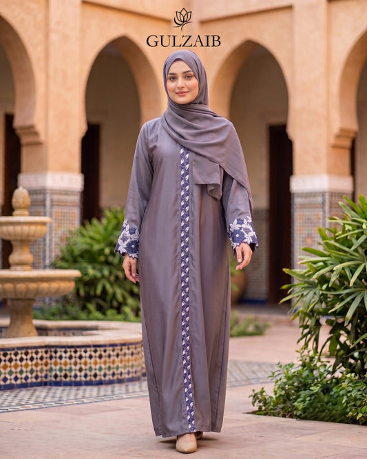 A woman wearing a long taupe-gray abaya with a matching hijab and patterned cuffs stands in front of an arched courtyard. The abaya features a vertical purple and white patterned stripe down the front and a matching purple patterned border.